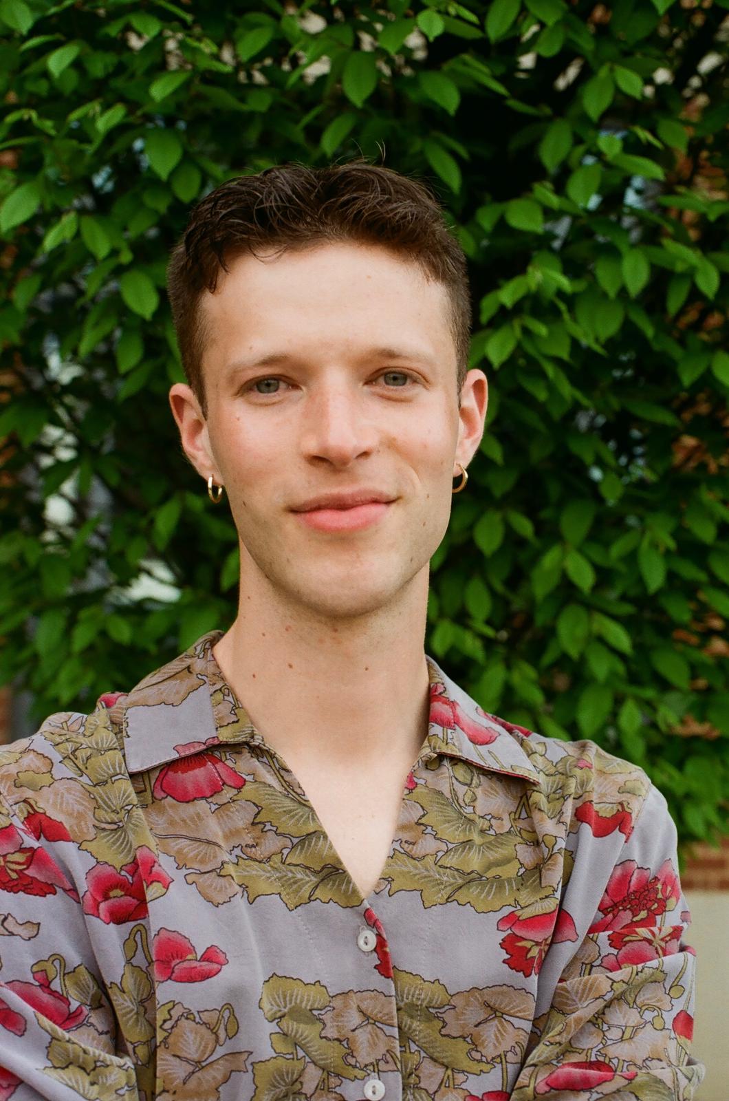 Isaiah stands in front of a green bush, wearing a purple collared shirt with red poppies on hit. He wears gold earrings and smiles gently.